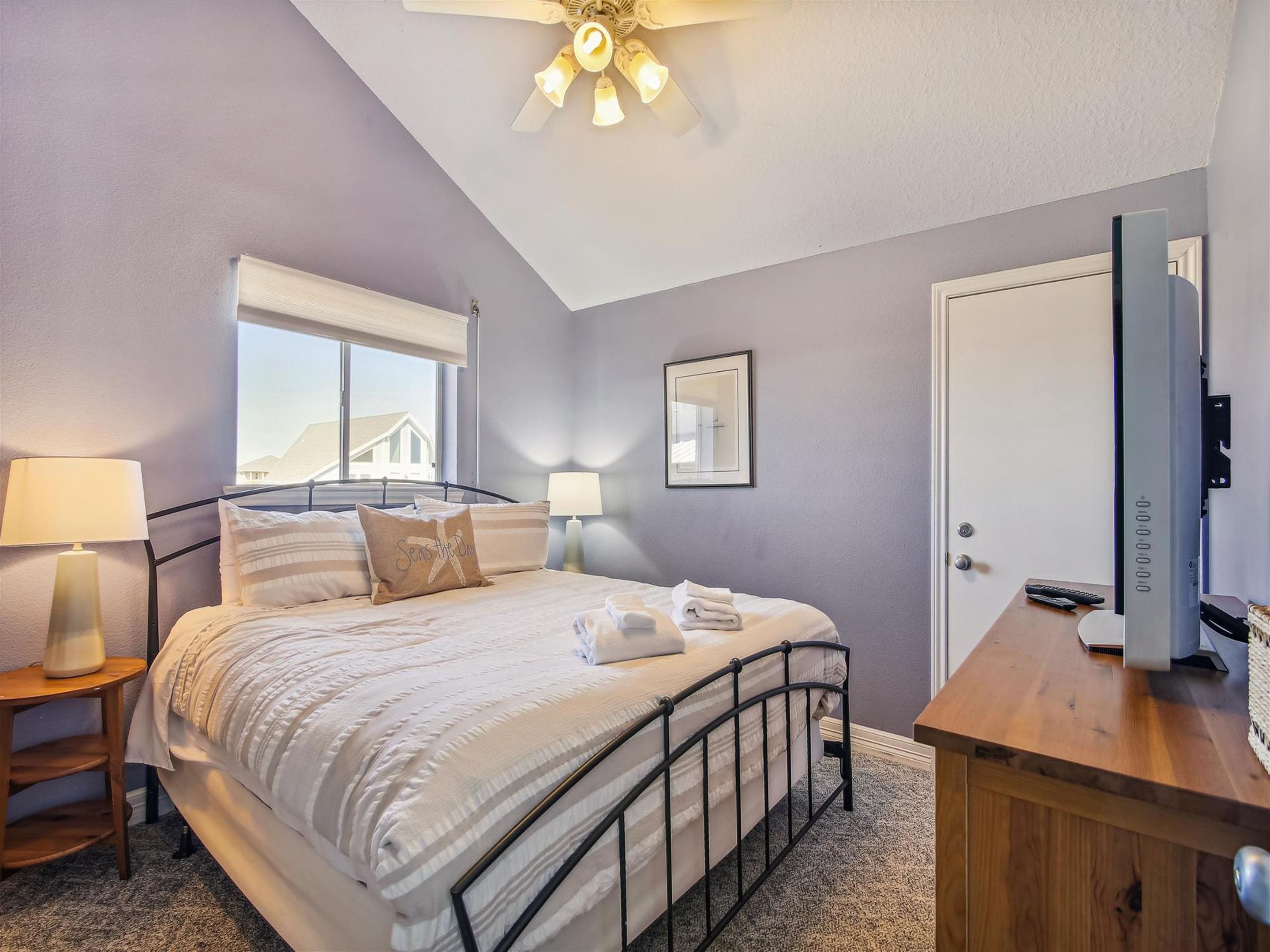 A bedroom with a bed, bedside lamps, a wooden dresser, a ceiling fan, a framed mirror, and a window with a view.