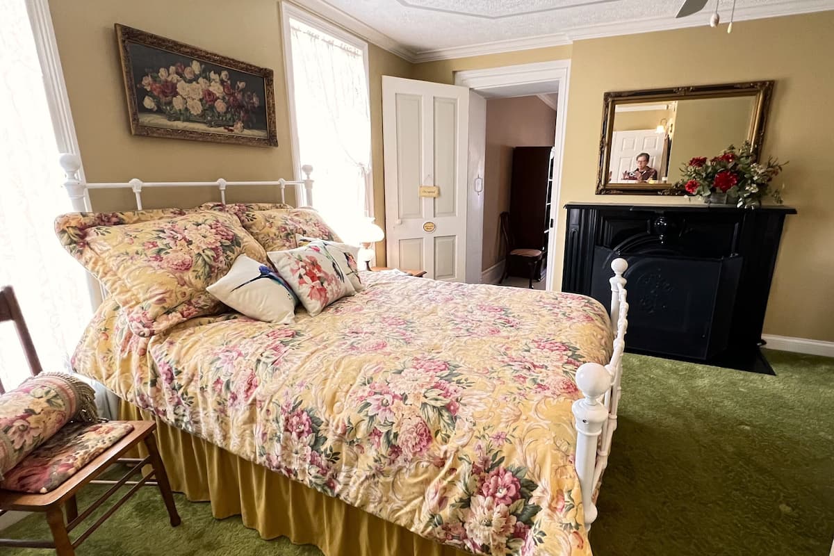 A bedroom with a floral bedspread, a wooden chair, a green carpet, a white bed frame, a ceiling fan, a fireplace, and a painting on the wall.