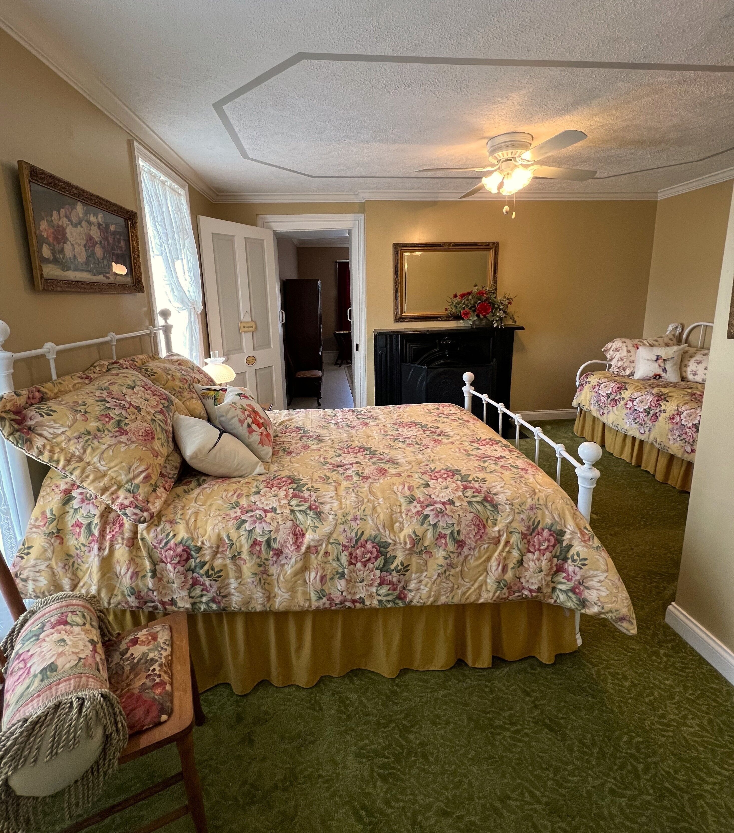 A bedroom with a floral bedspread, a wooden chair, a dresser, and a mirror on the wall.