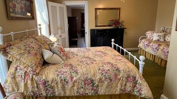A bedroom with a floral bedspread, a wooden chair, a dresser, and a mirror on the wall.