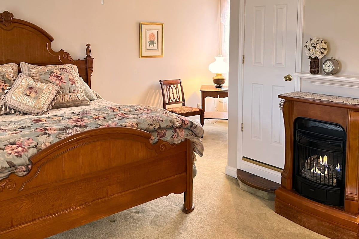 A bedroom with a wooden bed, a ceiling fan, a framed picture on the wall, a chair, a table with a lamp, and a fireplace.