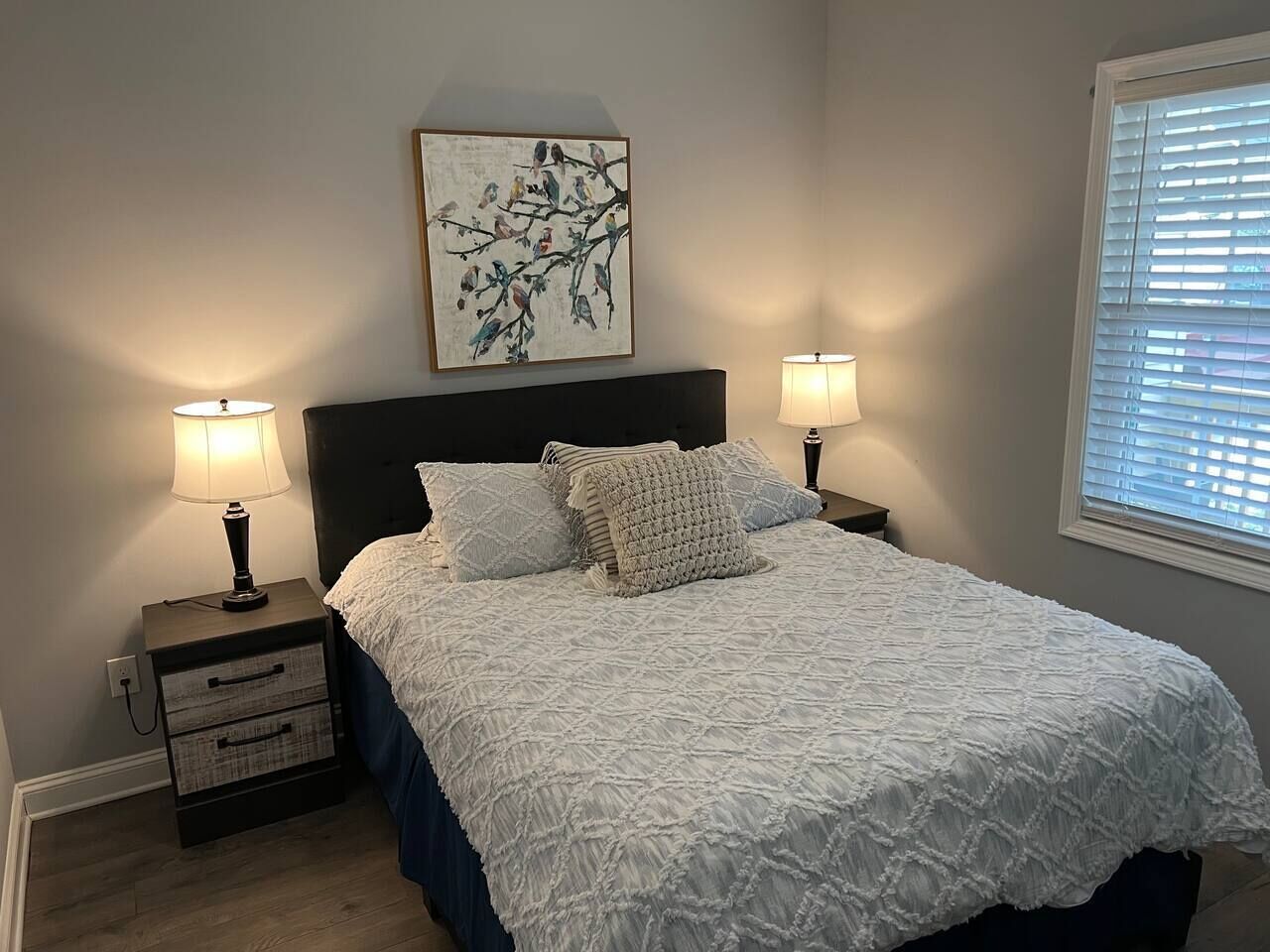 A bedroom with a bed, two bedside tables, a nightstand, a wall-mounted artwork, and a window with blinds.
