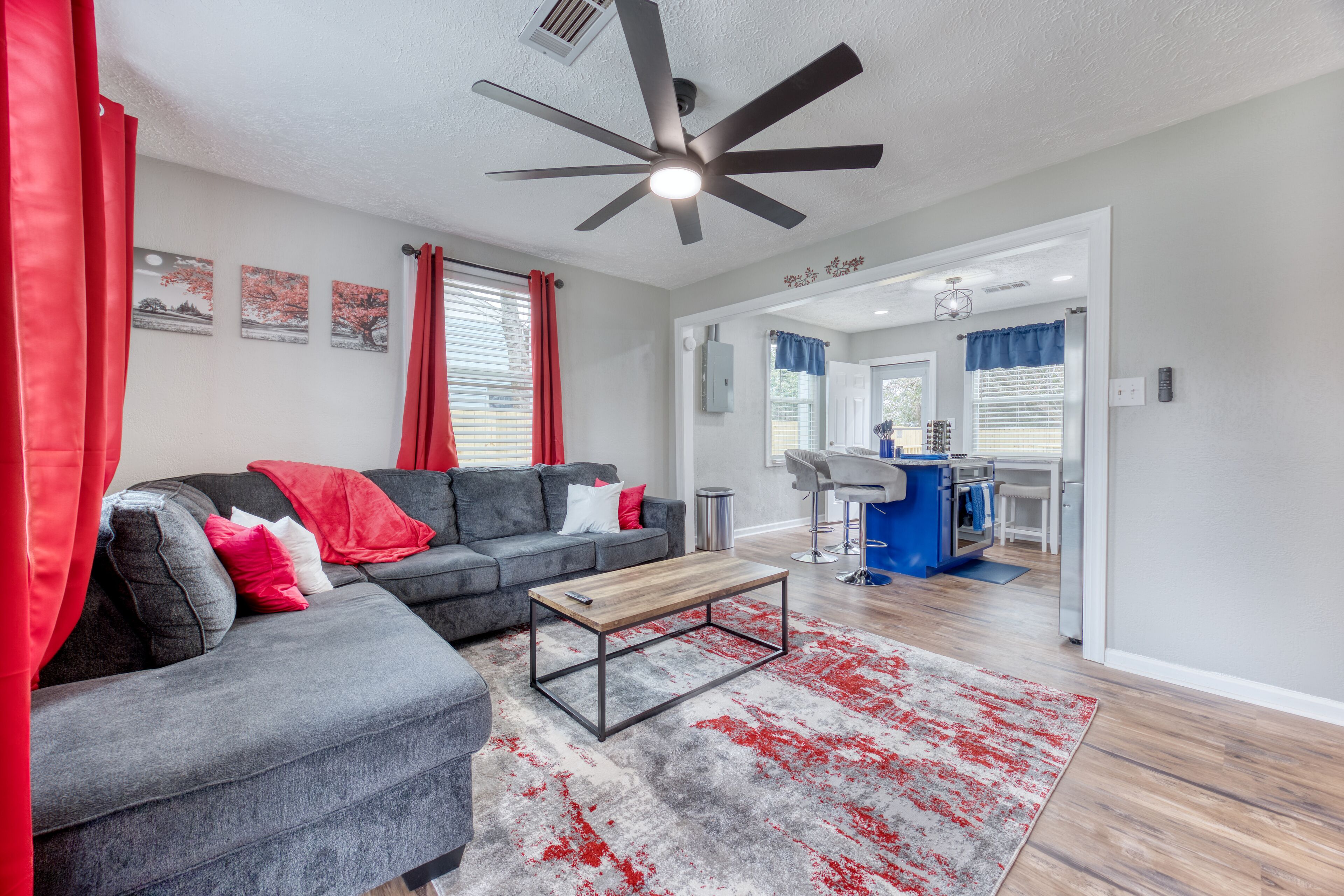 A modern living room with a grey sectional sofa, a wooden coffee table, and a red and grey rug. There is a ceiling fan, a kitchen area visible through an open doorway, and a dining area with bar stools.