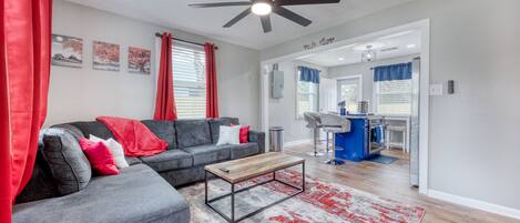 A modern living room with a grey sectional sofa, a wooden coffee table, and a red and grey rug. There is a ceiling fan, a kitchen area visible through an open doorway, and a dining area with bar stools.