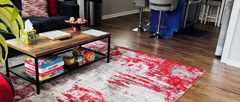 A living room with a sofa, coffee table, and a red and white rug. There is a kitchen area with a sink and a bar stool.