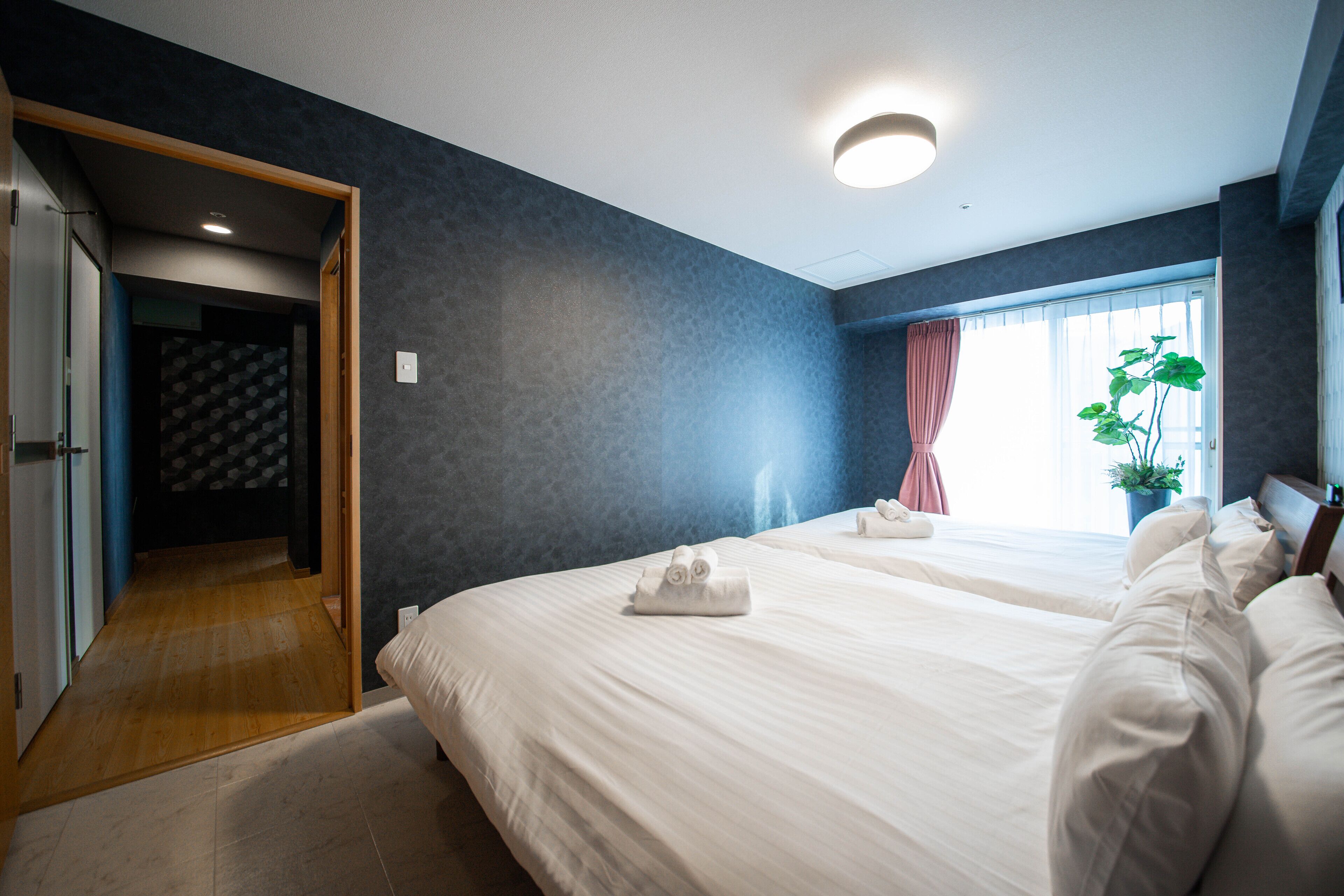 A hotel room with two beds, a wooden floor, a dark blue wall, a window with curtains, and a potted plant.
