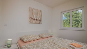 A bedroom with a bed, two pillows, a bedside lamp, a window with a view of trees, and a painting on the wall.