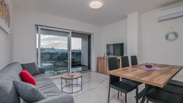 A modern living room with a grey sofa, a wooden dining table with chairs, a TV, and a sliding glass door leading to a balcony.