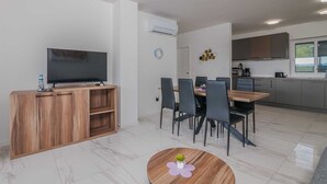 A modern living room with a flat-screen TV, wooden entertainment unit, dining area with chairs, and a kitchen with cabinets and a window.