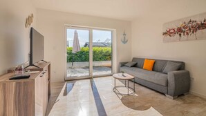 Ein modernes Wohnzimmer mit einem grauen Sofa, einem kleinen runden Tisch mit einer Pflanze, einem Fernseher auf einem Holzgestell und einer Glastür, die zu einem Außenbereich führt.