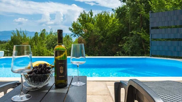 Une table au bord de la piscine, avec une bouteille de vin, deux verres et un bol de raisins.