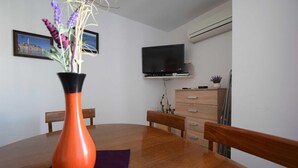Uma área de refeições com uma mesa de madeira, cadeiras, um vaso com flores, uma televisão em um suporte, uma cômoda e um aparelho de ar condicionado.