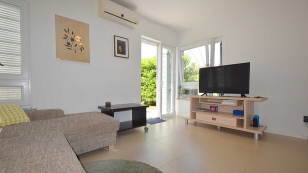 Una sala de estar moderna con un sofá, un televisor sobre un mueble de madera y un ventanal corredero que da a un balcón.