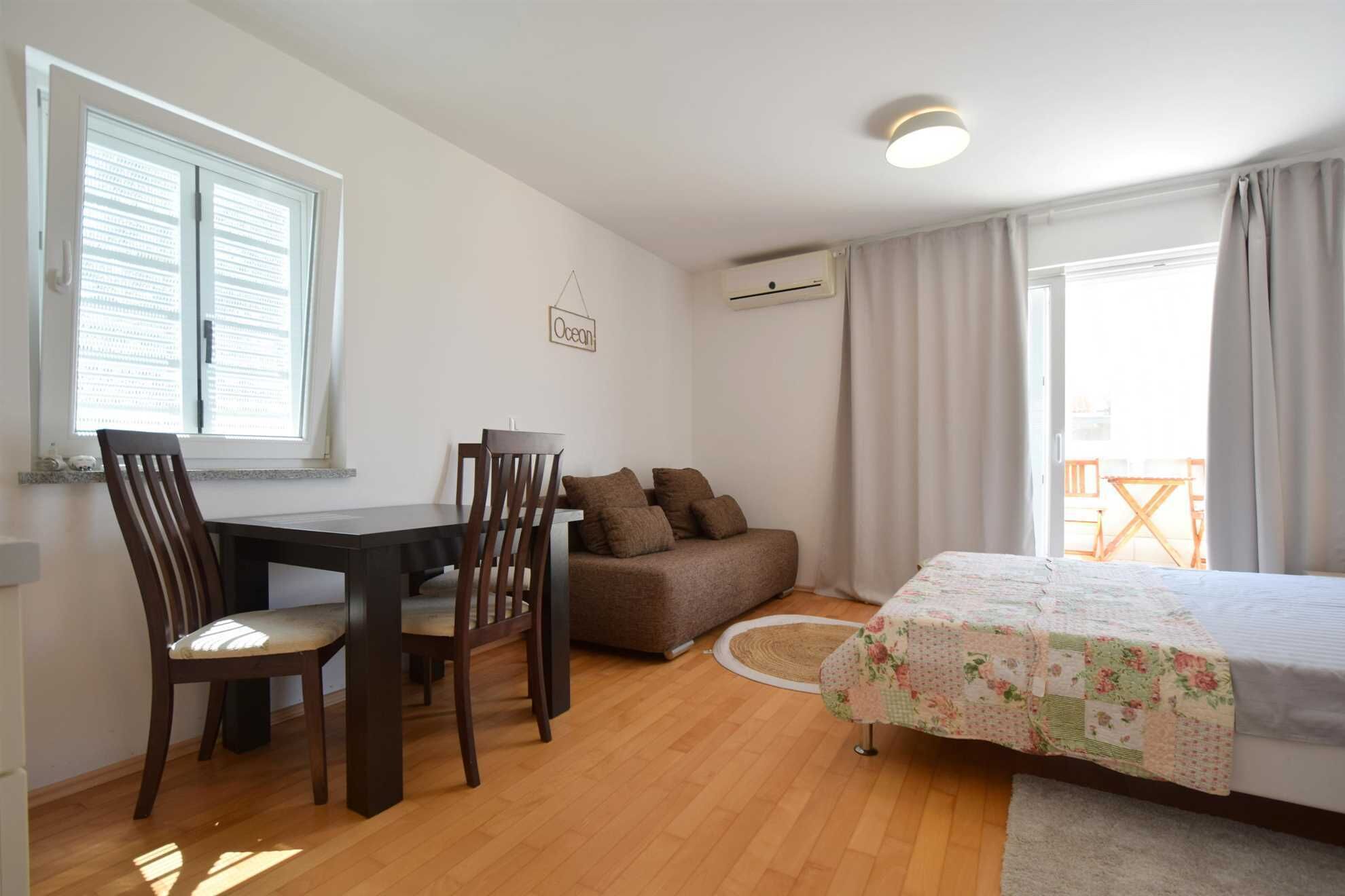 A compact bedroom with a bed, a sofa, a dining table with chairs, and a window with blinds.