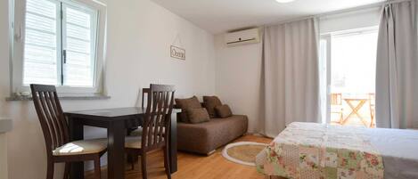 A compact bedroom with a bed, a sofa, a dining table with chairs, and a window with blinds.