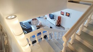 A staircase with white balusters leading to a landing. On the landing, there is a white sofa, a round table, and a floor lamp. Two framed pictures are on the wall.