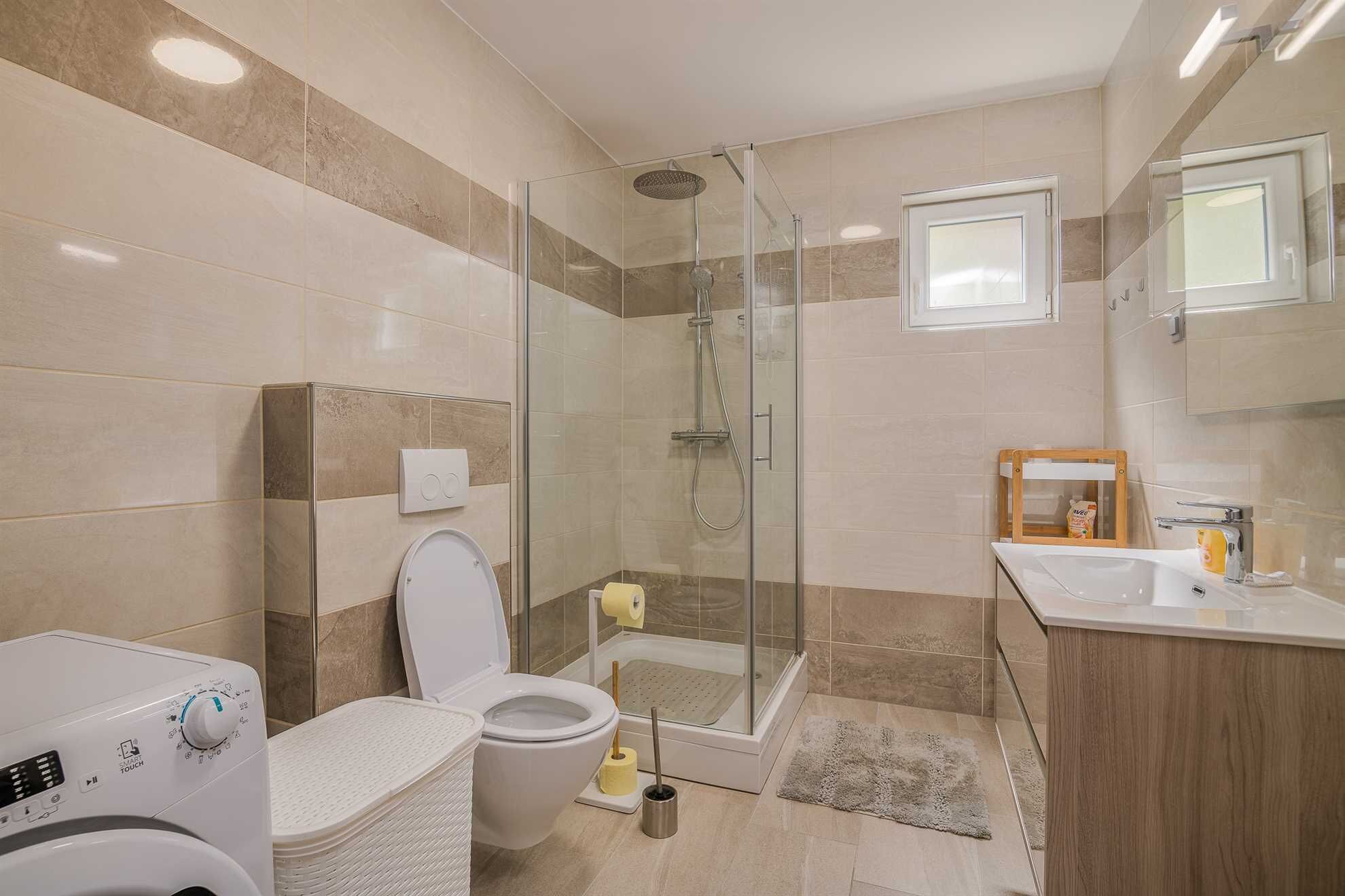 A modern bathroom with a glass-enclosed shower, a toilet, a washing machine, and a wooden vanity with a mirror.
