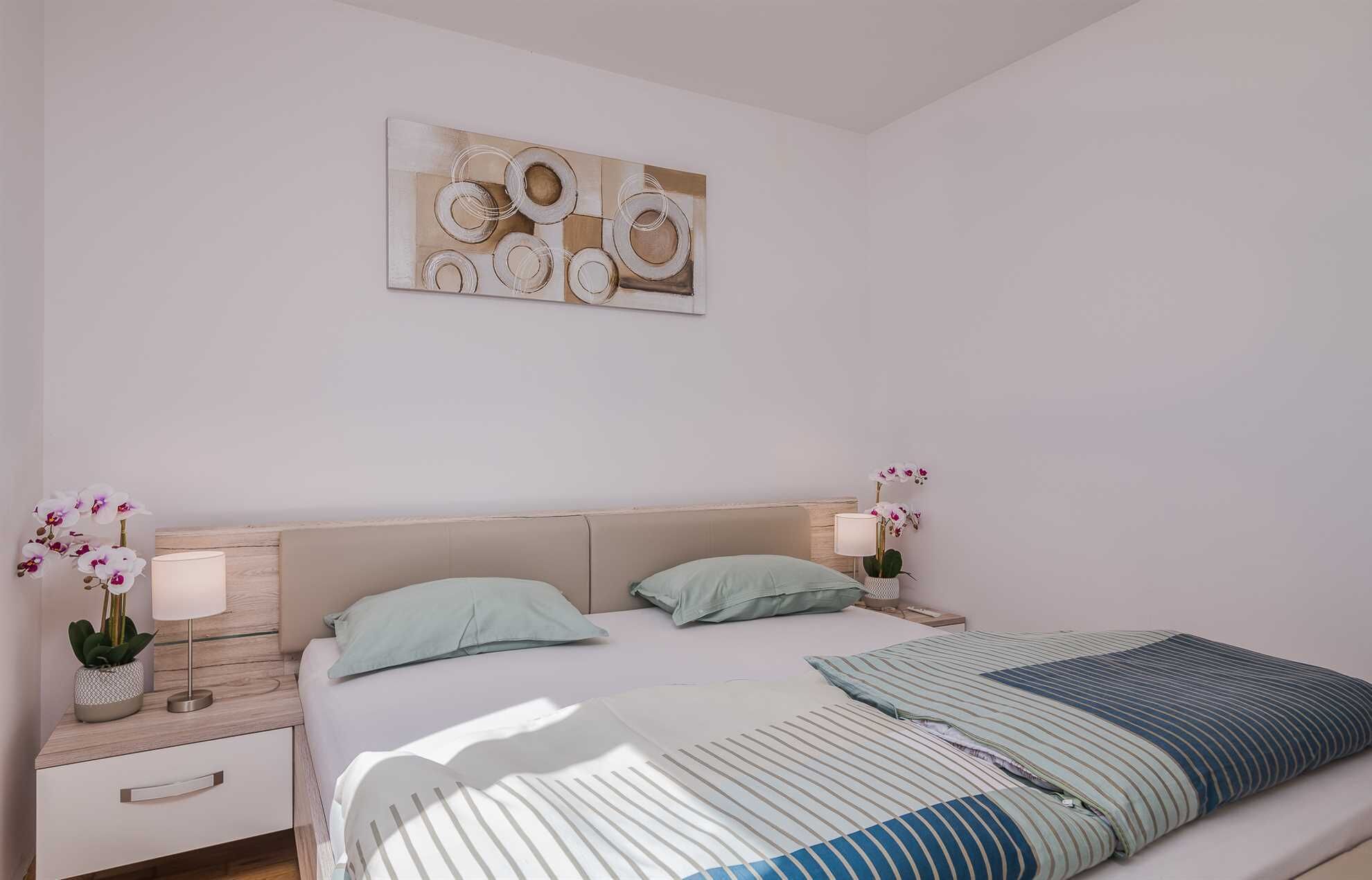 Une chambre à coucher comprenant un lit, deux tables de chevet, une table de nuit, une lampe, deux orchidées en pot et une œuvre d’art murale.