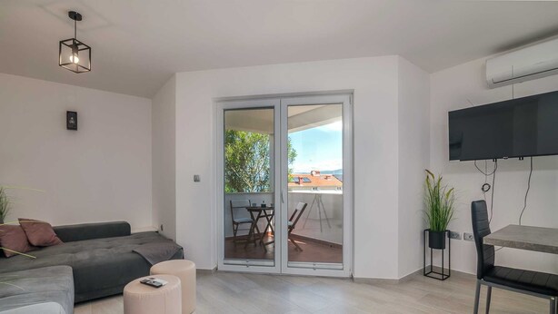 Un salon moderne avec un canapé gris, deux poufs, une table à manger avec des chaises, un téléviseur fixé au mur et une porte-fenêtre coulissante donnant sur un balcon.