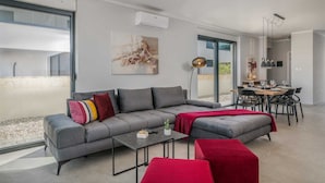 Ein modernes Wohnzimmer mit einem grauen Ecksofa, einem roten Hocker und einem Essbereich mit einem Holztisch und Stühlen.