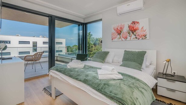 Ein modernes Schlafzimmer mit einem großen Bett, grüner Bettwäsche, Blick auf eine Stadtlandschaft und einem Balkon mit einem Stuhl.