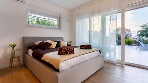 Ein modernes Schlafzimmer mit einem großen Bett, einem kleinen Nachttisch und Blick ins Freie durch eine Schiebetür.