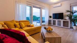 Ein modernes Wohnzimmer mit einem gelben Sofa, einem Flachbildfernseher, einem Holztisch mit Obst und großen Fenstern mit Blick ins Freie.