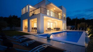 Ein modernes, zweistöckiges Haus mit Schwimmbad, Außenbereich mit Sitzgelegenheiten und einem Balkon mit Ziergeländer.