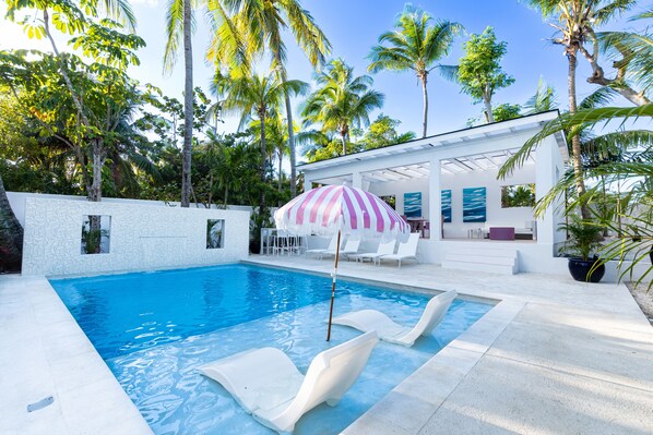 Piscina con sillas de descanso y un área de estar cubierta, rodeada de palmeras.