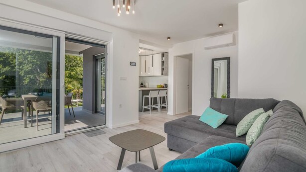 A modern living room with a grey sofa, teal cushions, a small coffee table, and a glass door leading to an outdoor area. - Apartment Manuela with private garden and hot whirlpool (Njivice)