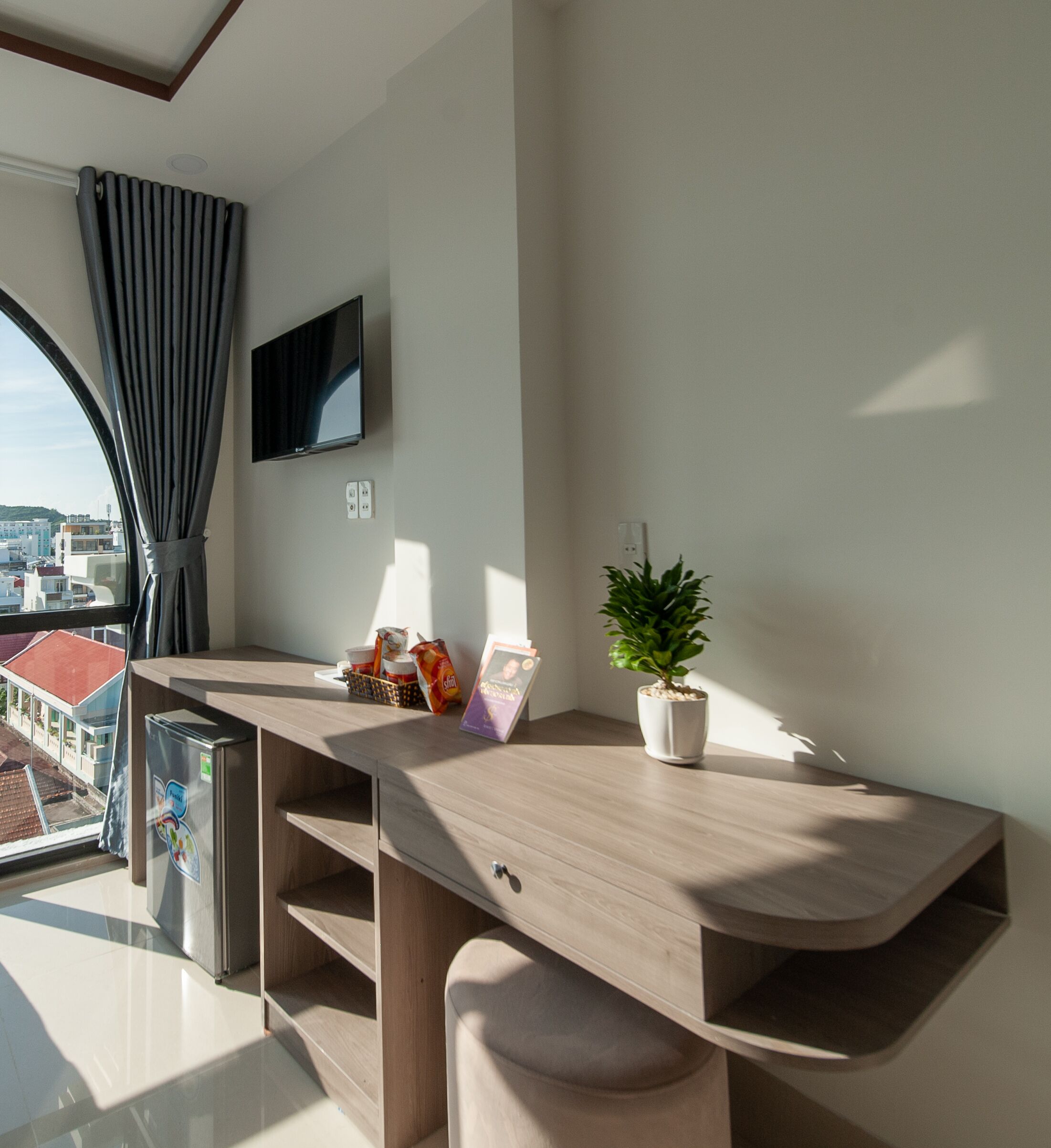 A modern room with a wooden desk, a flat-screen TV mounted on the wall, a potted plant, and a view of the city through a large window.
