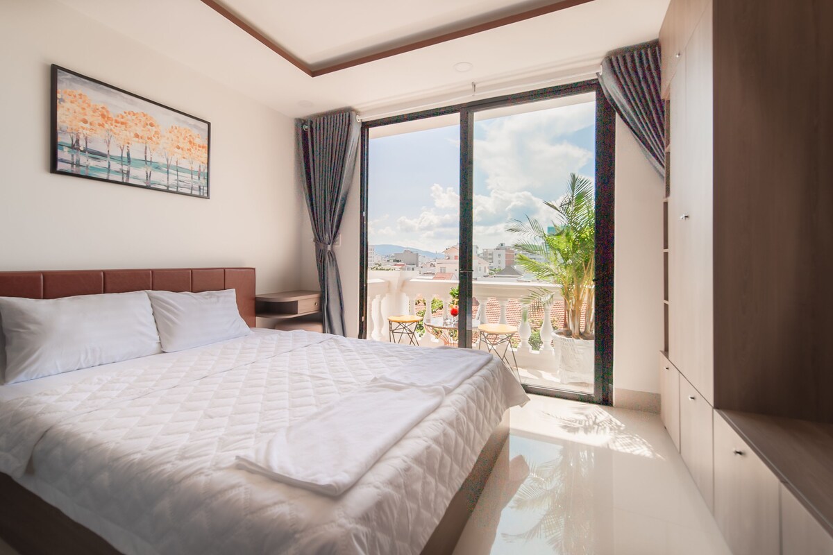 Un dormitorio con cama, mesita de noche, un cuadro, ventana con cortinas y vista a la ciudad.