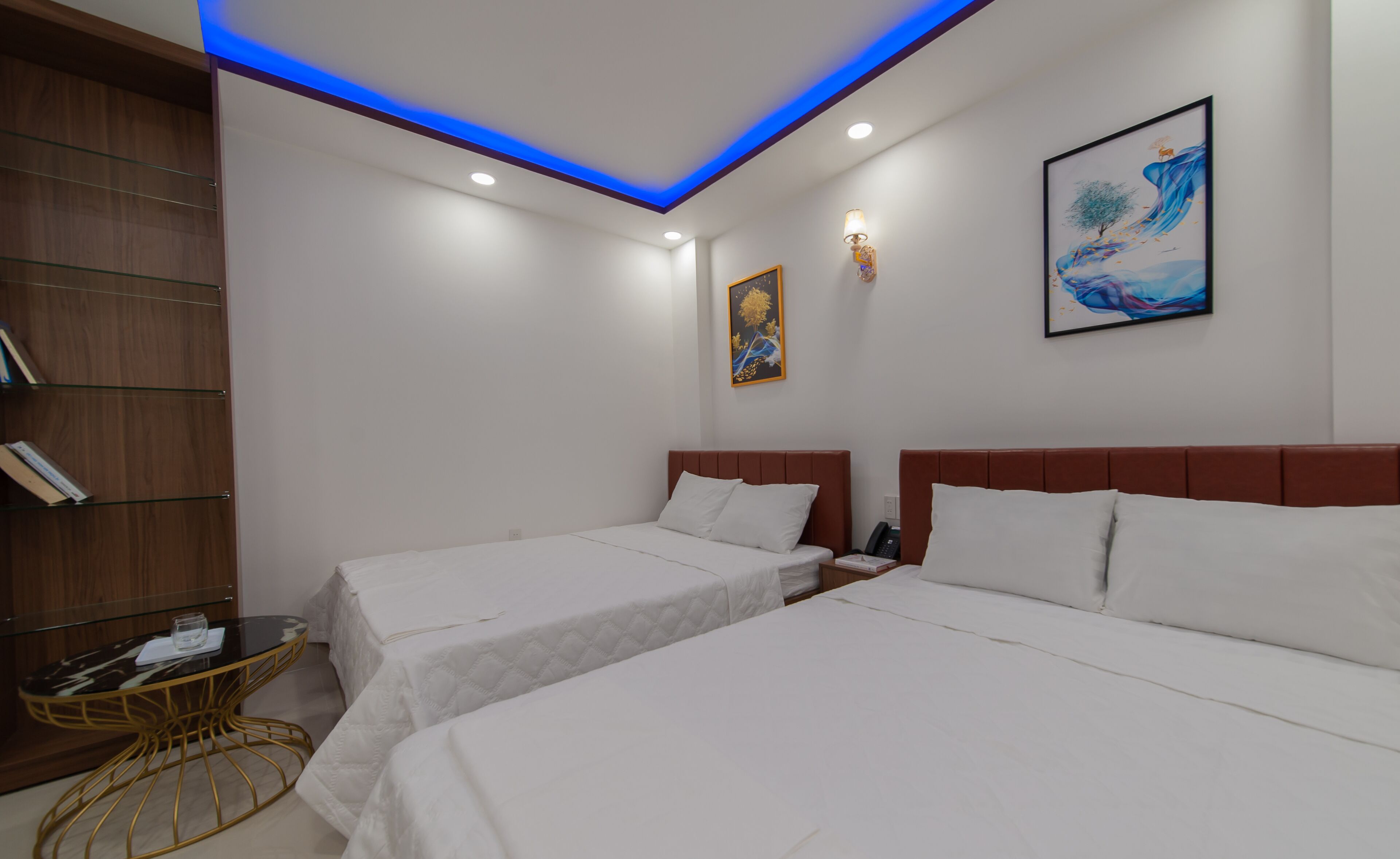 Habitación de hotel con dos camas, una mesita, una estantería y un cuadro en la pared.