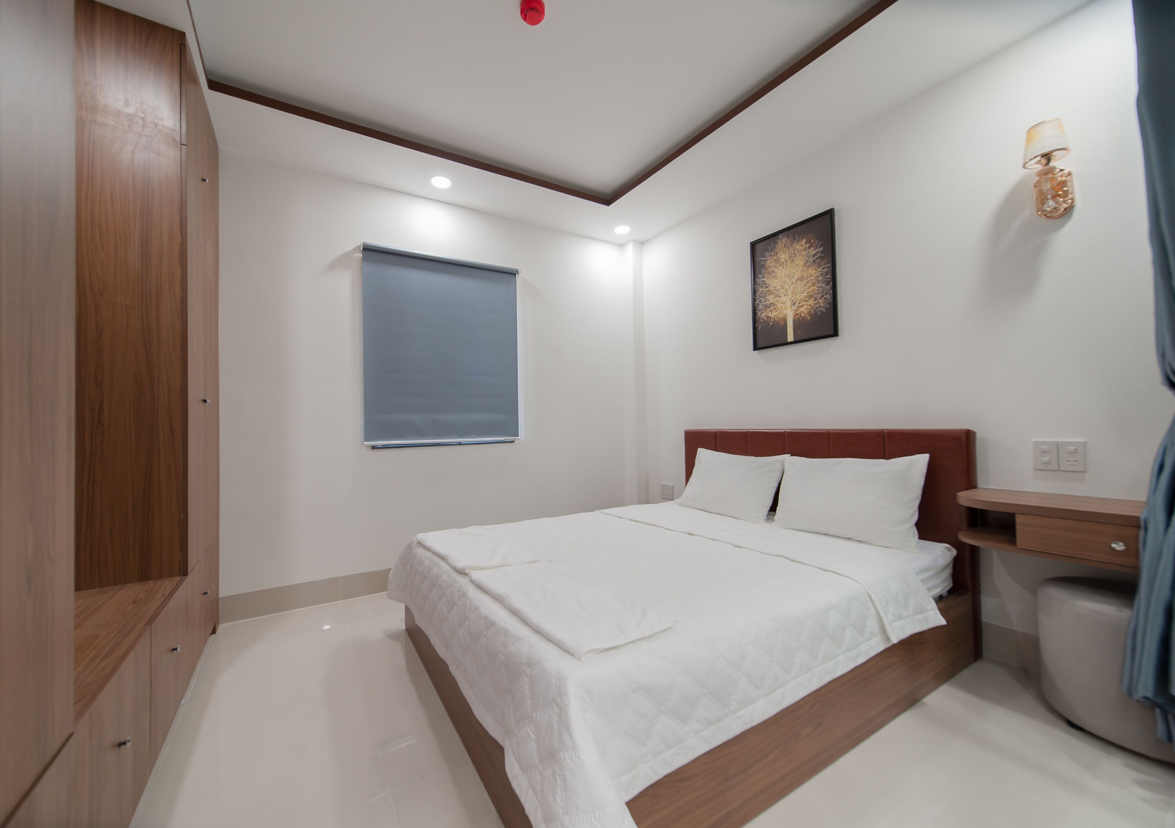 A bedroom with a bed, a nightstand, a wall-mounted lamp, a window with blinds, and a ceiling light.