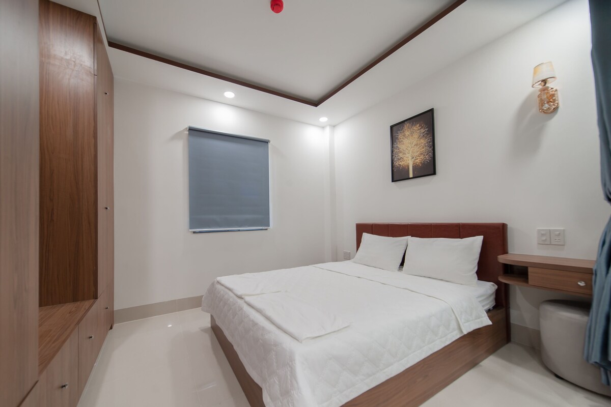 Un dormitorio con cama, mesita de noche, lámpara de pared, ventana con persianas y luz de techo.