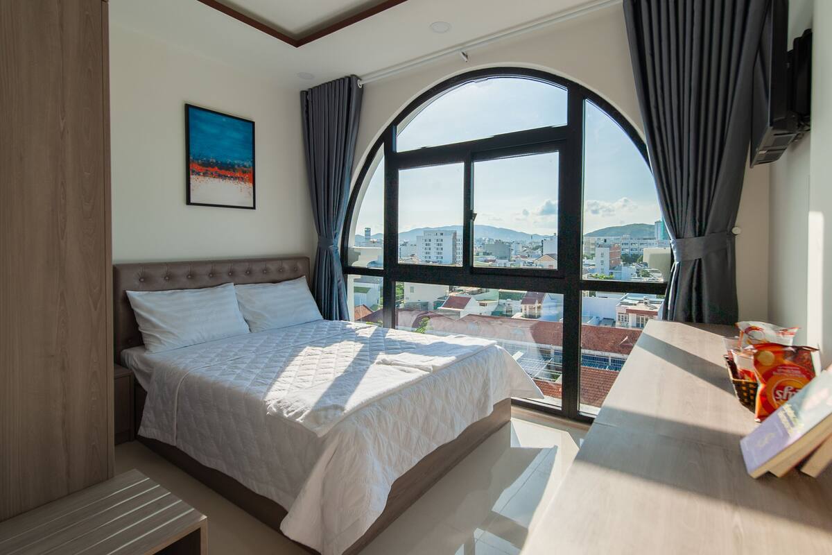 Una camera da letto con un'ampia finestra, un letto, un comodino e vista sulla città.