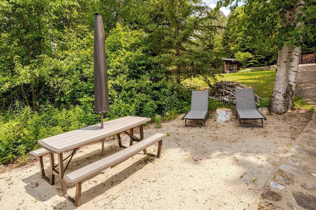 Una mesa de picnic con banco y dos sillas reclinables en una zona arenosa rodeada de vegetación.