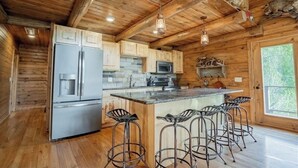 A rustic cabin-style kitchen with wooden walls, a central island with bar stools, stainless steel appliances, and a large window. - Buffalo County Log Cabin w Guesthouse and pool (Alma)