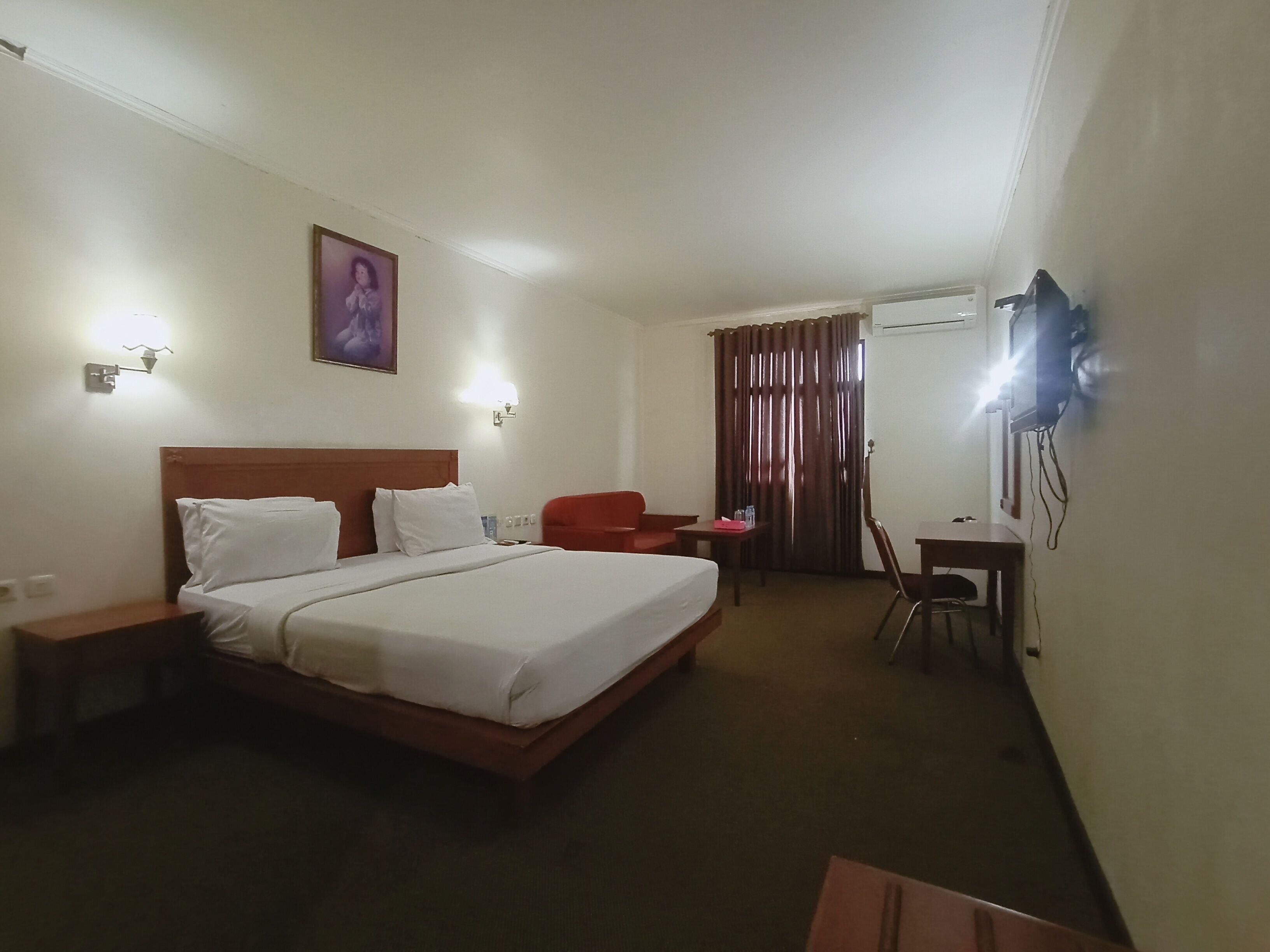 A hotel room with a bed, a desk, a chair, a red armchair, a TV mounted on the wall, and a curtain.