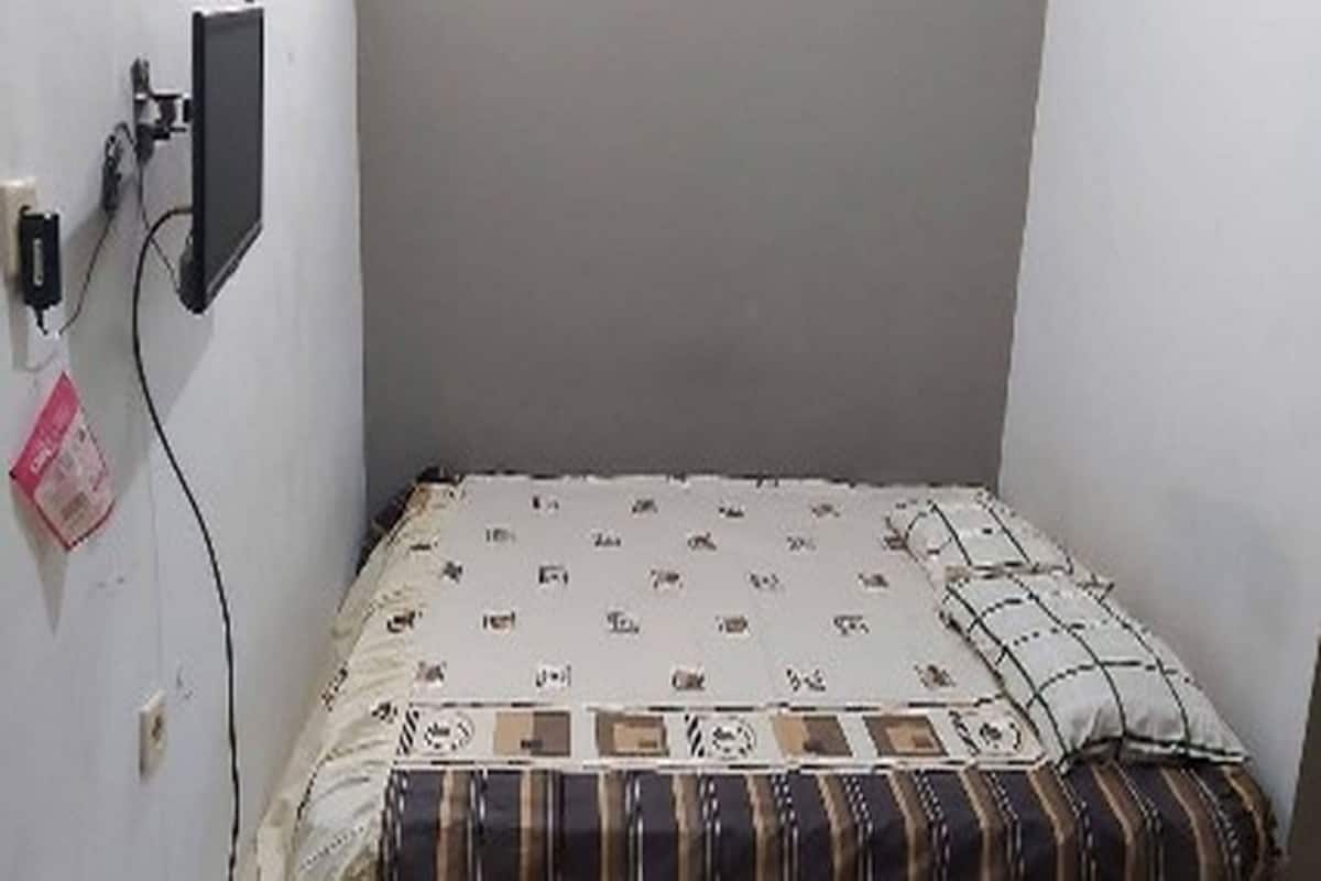 A small, sparsely furnished room with a bed, a power outlet, and a wall-mounted television.
