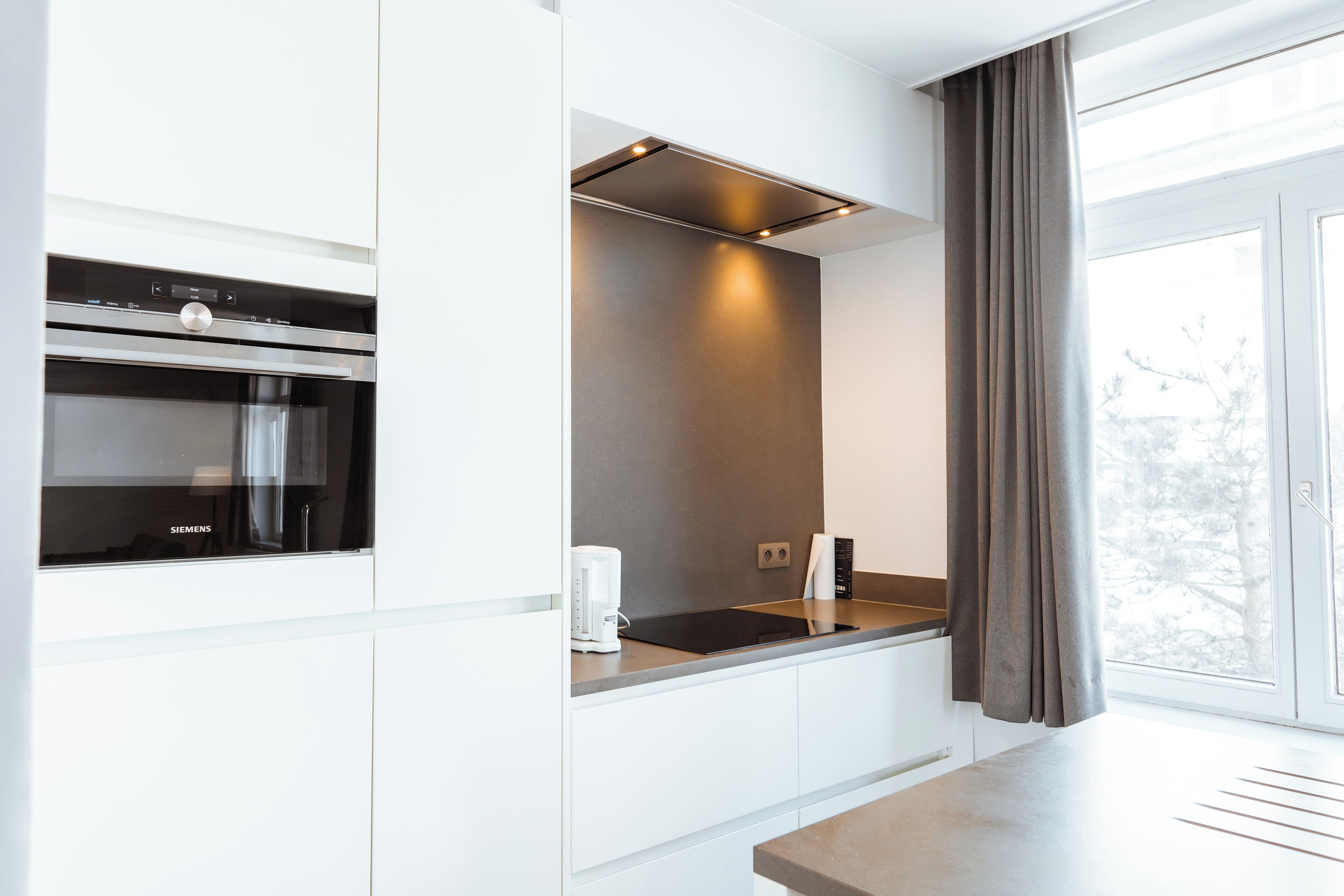 Una cocina moderna con horno empotrado, cafetera y ventana con cortinas.