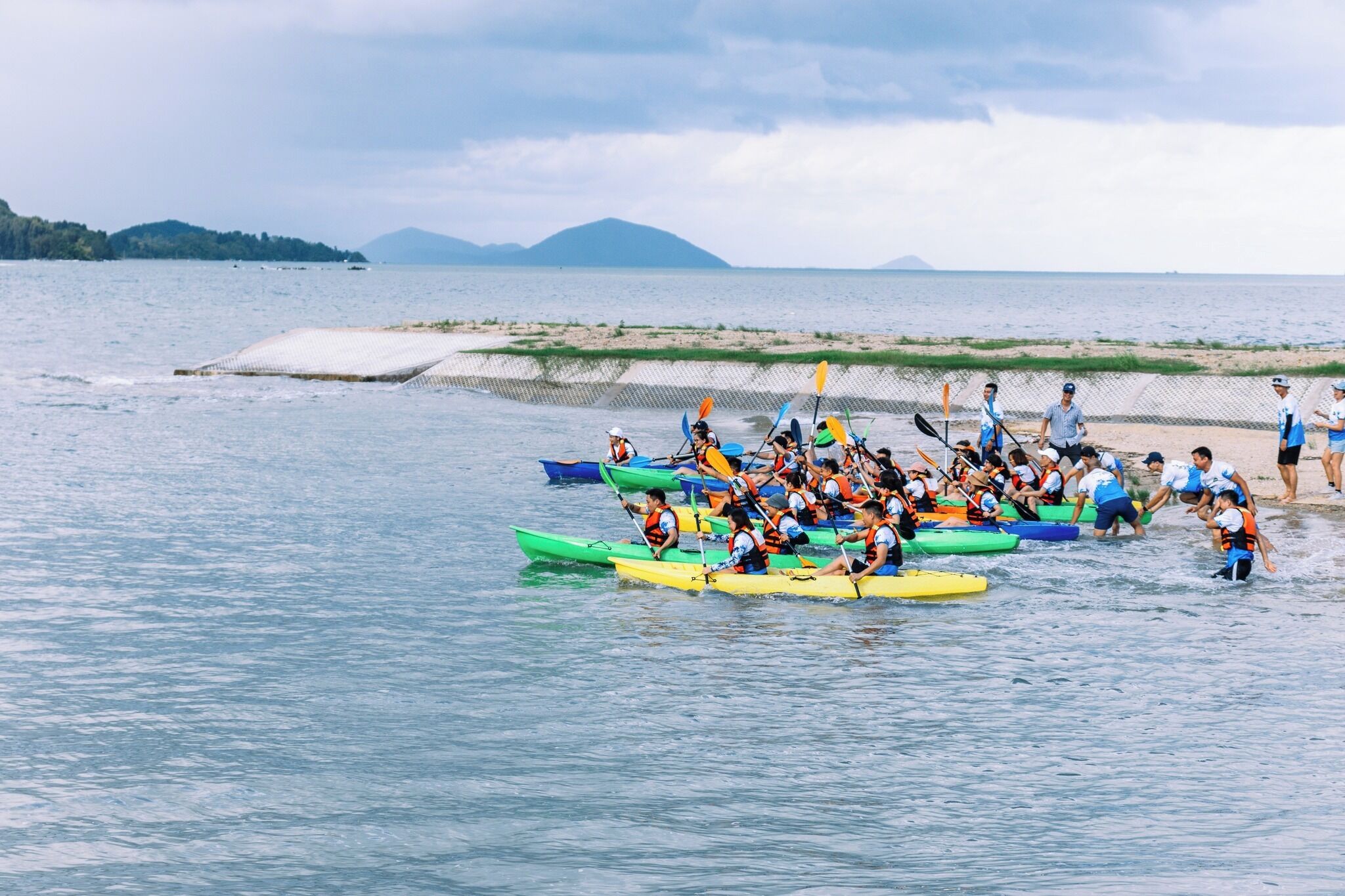 Een groep mensen in kleurrijke kajaks op kalm water, met een zandstrand en bergen in de verte.