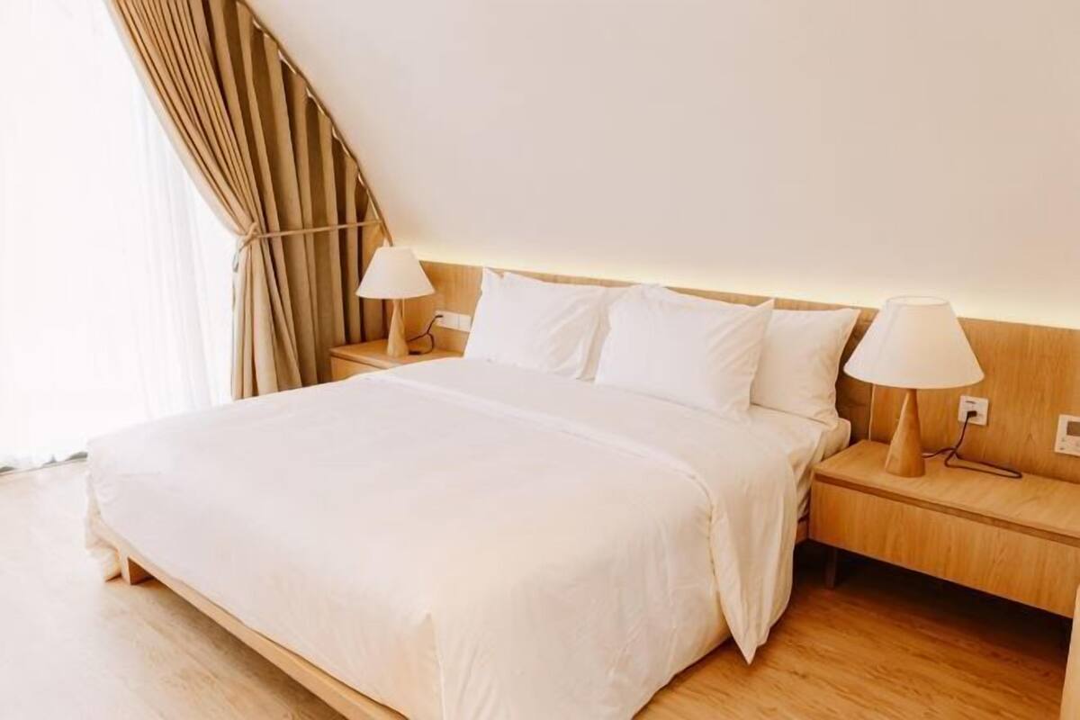 A hotel room with a large bed, two bedside tables with lamps, wooden flooring, and a window with curtains.