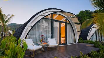Un edificio moderno de forma abovedada con amplias ventanas de vidrio, una terraza de madera y mobiliario blanco.
