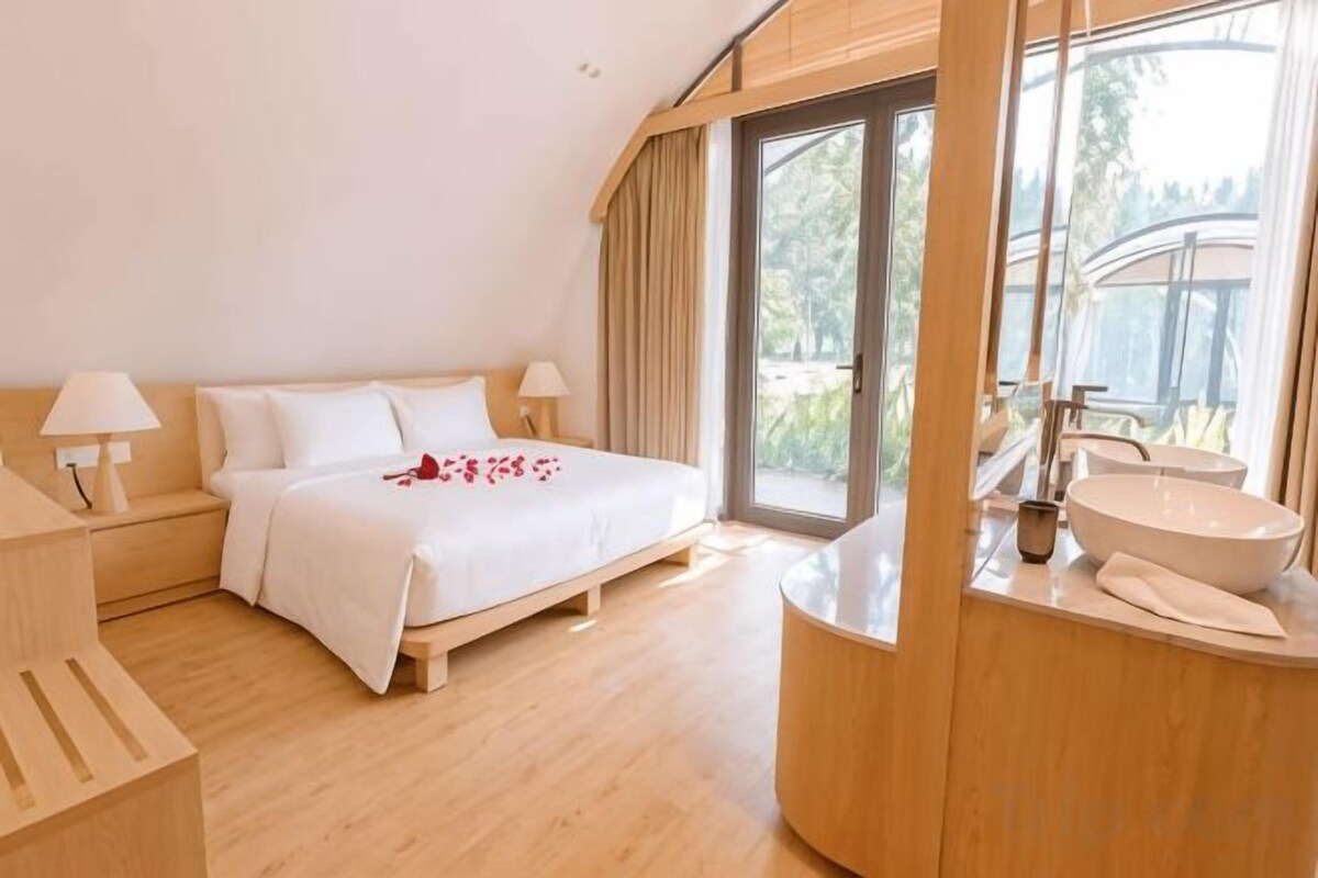 A modern bedroom with a bed, bedside tables, a large window, and a bathroom with a sink and mirror.