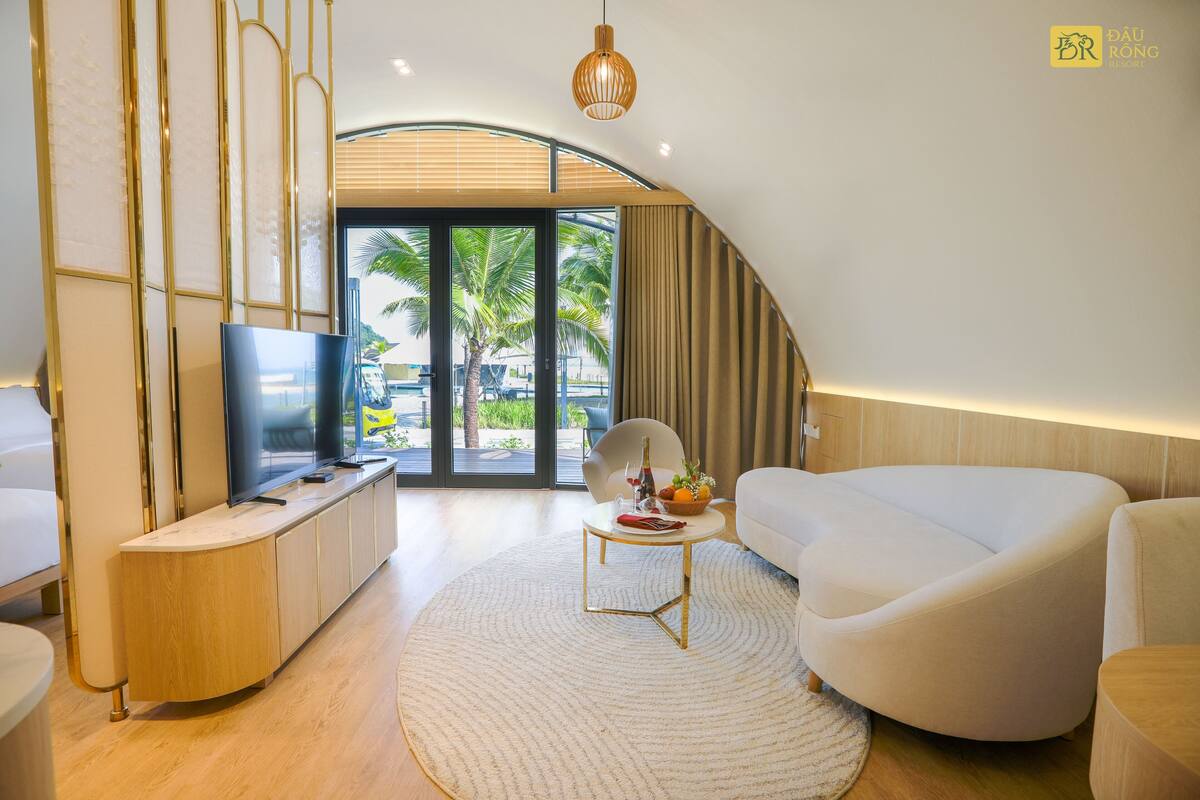 A modern living room with a curved ceiling, a flat-screen TV, a white sofa, a round coffee table, and a view of palm trees through large glass doors.