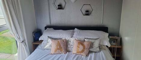 A bedroom with a bed, white bedding, a wooden headboard, two hexagonal wall sconces, and a small bedside table with a picture frame.