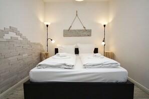 A double bed with white linens and towels, two bedside tables with lamps, a brick accent wall, and a rustic wooden headboard.
