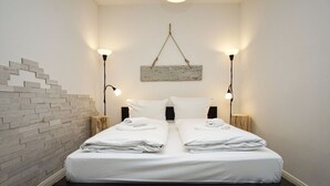 Ein Doppelbett mit weißen Leinen und Handtüchern, zwei Nachttische mit Lampen, eine Wand mit Ziegeln als Blickfang und ein rustikales Holz-Kopfteil.