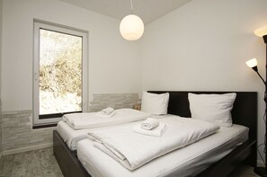 Une chambre à coucher moderne avec une grande fenêtre, un lit recouvert de draps blancs soigneusement pliés et un design minimaliste.